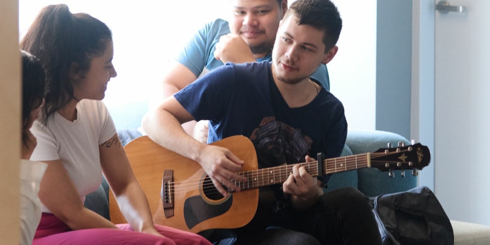 Image of a group of students sitting down. A person in the middle is playing guitar