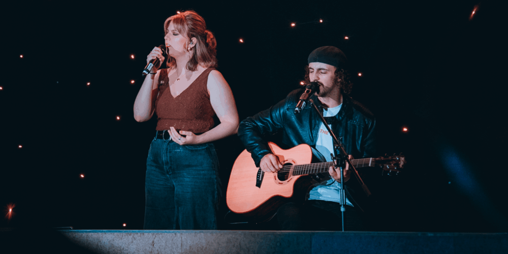 A female singer performing next to a male student playing acoustic guitar.